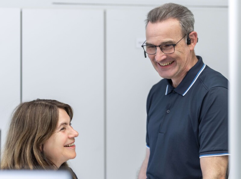 Auf dem Bild ist links eine Dame sitzend und rechts ein Herr stehend mit Brille und Headset zu sehen. Die Personen sind sich zugewandt und lachen.