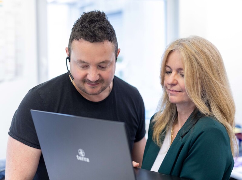 Zwei Mitarbeiter der QUACERT, eine Dame mit offenem Laptop und ein junger Mann, welcher rechts von ihr steht und ebenfalls in den Laptop sieht. Der junge Mann trägt ein Headset.