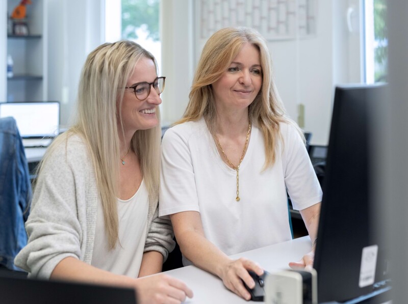 Auf diesem Bild sind zwei junge blonde Frauen zu sehen welche am Schreibtisch sitzen und gemeinsam auf einen Monitor schauen und dabei lächeln. Die linke jüngere Dame trägt eine Brille.